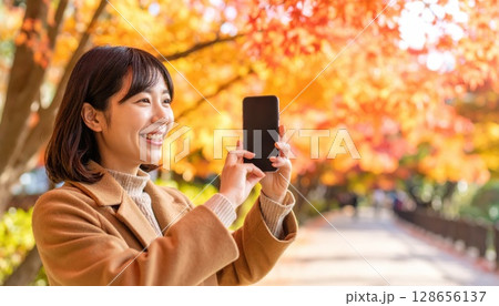 《AI画像》秋の紅葉をスマホで撮影する若い女性 128656137