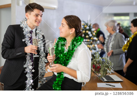 Two joyful male and female coworkers with tinsels and glasses of champagne during corporate New Year party in office Two joyful male and female coworkers with tinsels and glasses of champagne during corporate New Year party in office 128656386