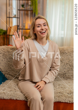 Adult woman smiling and waving hand at home, greeting someone with joy and cheerful energy on sofa Adult woman smiling and waving hand at home, greeting someone with joy and cheerful energy on sofa 128656520