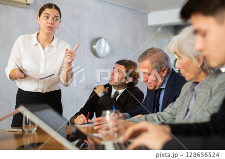 Clever young female making report while office workers of different ages using notebooks at table in office Clever young female making report while office workers of different ages using notebooks at table in office 128656524