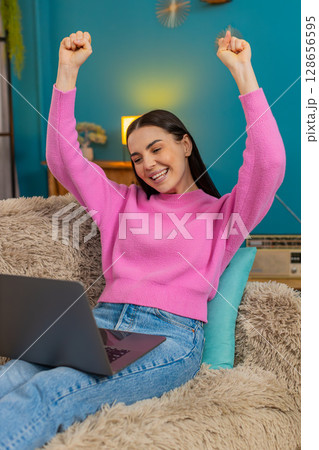 Young woman on sofa raising hands up joyfully celebrating successful finished project on laptop 128656595