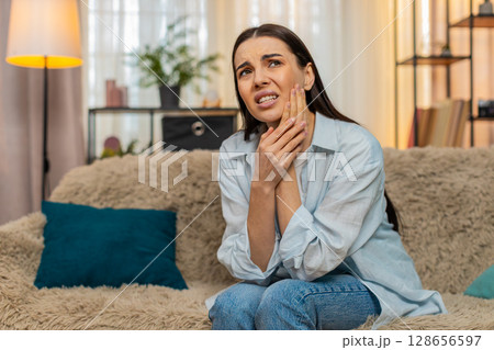 Young woman sitting on sofa holding cheek suffering from strong toothache feeling pain uncomfortable Young woman sitting on sofa holding cheek suffering from strong toothache feeling pain uncomfortable 128656597