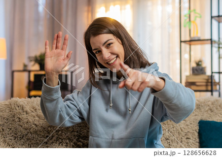 Young Caucasian woman girl on sofa joyfully exclaims Hey you and waves hand palm in hello gesture Young Caucasian woman girl on sofa joyfully exclaims Hey you and waves hand palm in hello gesture 128656628