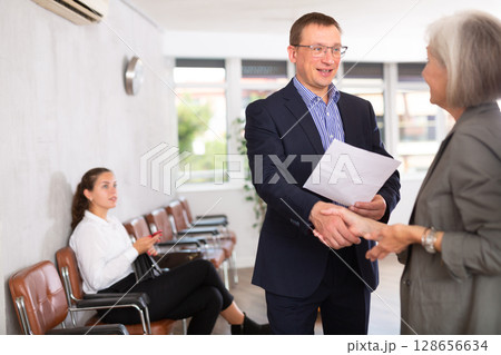 Senior woman makes deal, shaking hands with middle-aged man colleague while standing in office 128656634