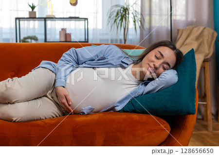Smiling young pregnant woman stroking big belly while sleeping on comfortable sofa dreaming at home 128656650