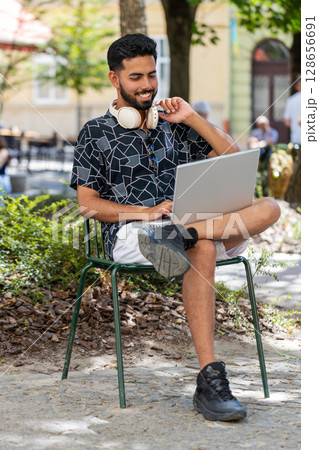 Indian man in headphones working online distant job with laptop browsing website chatting outdoors Indian man in headphones working online distant job with laptop browsing website chatting outdoors 128656691