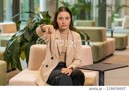 Portrait of upset Caucasian businesswoman sitting in office lobby showing thumb down sign gesture 128656697
