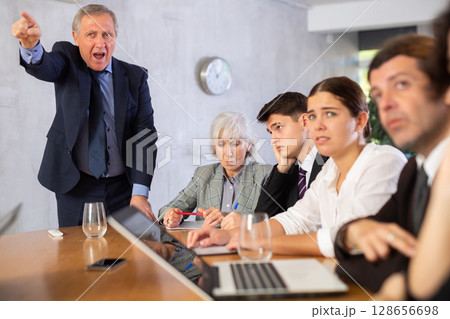 Outraged old manager getting mad at young adult mature office workers sitting with reports and laptops at table in meeting room Outraged old manager getting mad at young adult mature office workers sitting with reports and laptops at table in meeting room 128656698