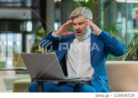 Sad businessman using laptop loses becoming shocked sudden lottery results in modern hotel lobby 128656729