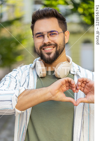 Smiling Caucasian young man makes symbol of love, showing heart sign to camera on city town street Smiling Caucasian young man makes symbol of love, showing heart sign to camera on city town street 128656741
