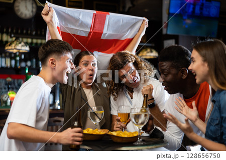 Males and females with England flag clinking glasses with beer on party in bar Males and females with England flag clinking glasses with beer on party in bar 128656750