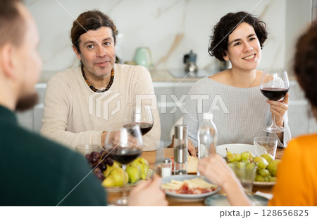 Group of joyful individuals are celebrating a holiday, enjoying drinks and conversation around table in their home 128656825
