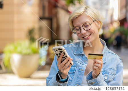 Happy Caucasian woman using credit card smartphone while purchases online shopping on city street Happy Caucasian woman using credit card smartphone while purchases online shopping on city street 128657131