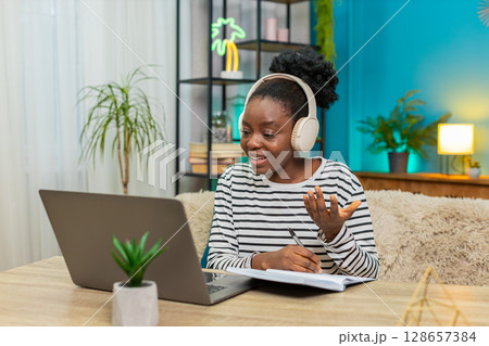 African American woman studying online headphones notes laptop sofa home table indoors education 128657384
