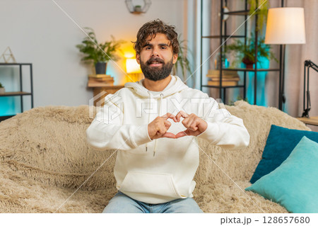Happy Indian young man at home living room couch makes symbol of love, showing heart sign to camera 128657680