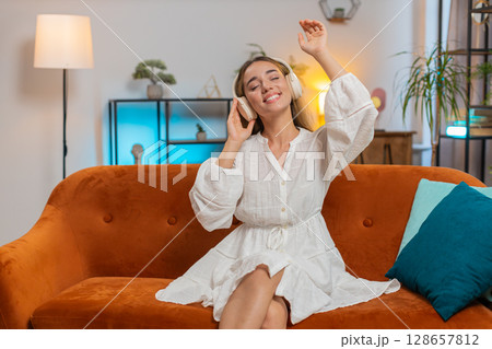 Joyful young Caucasian woman listening to music through wireless headphones sitting on sofa couch 128657812
