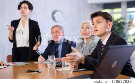 Concentrated young adult old office workers sitting around the table and looking at screen while Latin female using projector remote control in office 128657820
