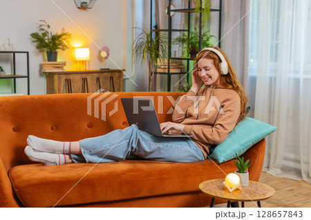 Happy relaxed Caucasian young woman in wireless headphones listening music using laptop on sofa 128657843