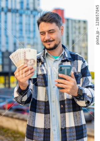 Happy rich mature business man using smartphone and counting money hryvnias on downtown city street 128657845