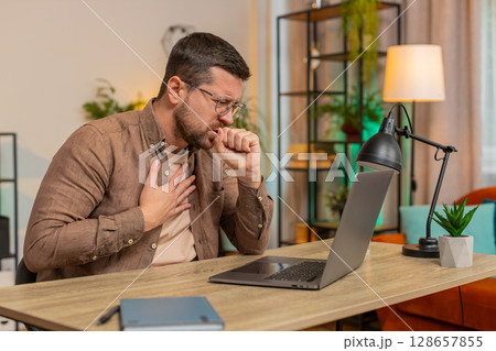 Sick man freelancer feeling unwell suffering from sore throat touches chest while coughing at home Sick man freelancer feeling unwell suffering from sore throat touches chest while coughing at home 128657855