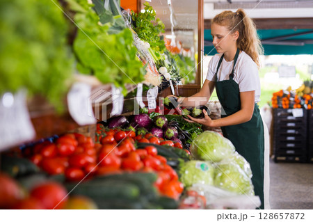 Young girl seller checking organic eggplants Young girl seller checking organic eggplants 128657872