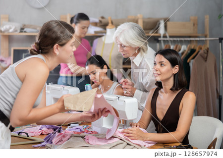 Girl stitching at machine and talking to groupmate in sewing workshop 128657974