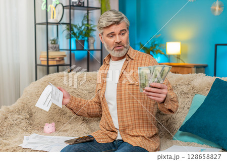 Mature man on sofa using smartphone calculator, counting cash and bills, feeling exhausted at home Mature man on sofa using smartphone calculator, counting cash and bills, feeling exhausted at home 128658827