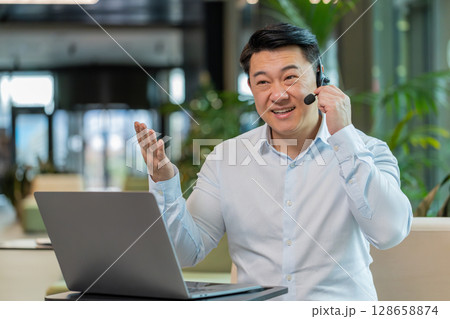 Mature businessman with laptop wearing headset support service talking with client in office lobby 128658874