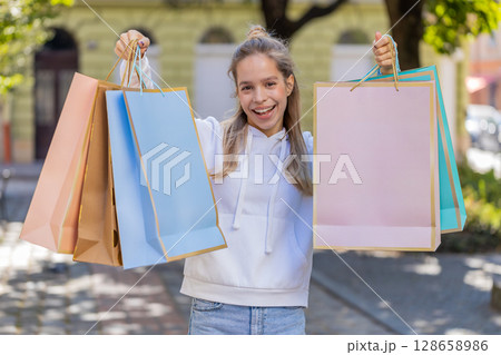 Happy smiling young Caucasian woman shopaholic carrying shopping bags and walking on city street 128658986
