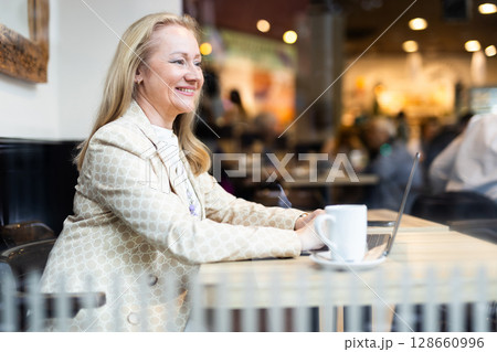 Business lunch - businesswoman sits at table in cafe by window with a laptop and drinks coffee. View through window Business lunch - businesswoman sits at table in cafe by window with a laptop and drinks coffee. View through window 128660996