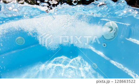 Underwater photo of a powerful water jet in a swimming pool, creating bubbles and turbulence near the pool wall with clear blue water and light reflections. 128663522