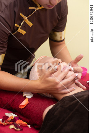 a masseuse in a cosmetology salon makes a Thai massage on the face of a young girl a masseuse in a cosmetology salon makes a Thai massage on the face of a young girl 128671941