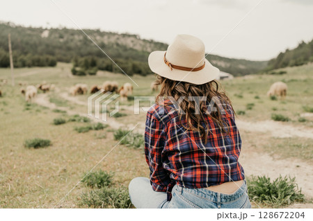 A woman in a plaid shirt and hat is sitting in a field with a herd of sheep A woman in a plaid shirt and hat is sitting in a field with a herd of sheep 128672204