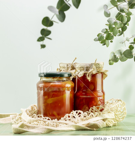 Probiotic foods. Eco friendly still life with pickled or fermented vegetables. Lecho and ajvar in glass jars on mesh bag on blue background with eucalyptus sprigs. Home food preserving or canning Probiotic foods. Eco friendly still life with pickled or fermented vegetables. Lecho and ajvar in glass jars on mesh bag on blue background with eucalyptus sprigs. Home food preserving or canning 128674237