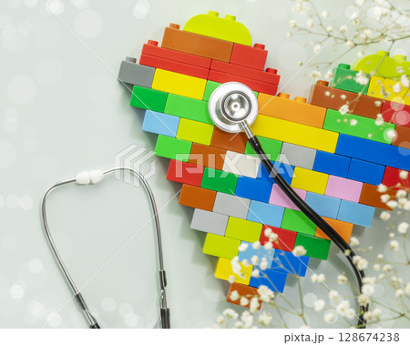 Multicolored kid bricks heart shape with stethoscope on blue background with white flowers. Health care, pediatric, love, and family insurance concept, world heart day. Flat lay 128674238