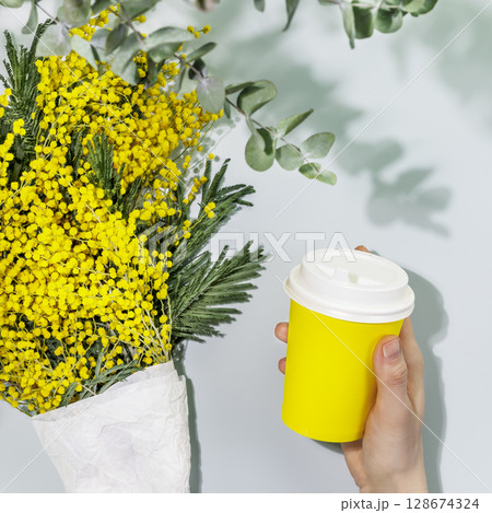 Coffee to go. Spring life style with bouquet of mimosa flowers and female hand holds disposable cardboard cup with coffee on blue background. Takeaway coffee concept. Mockup, flat lay Coffee to go. Spring life style with bouquet of mimosa flowers and female hand holds disposable cardboard cup with coffee on blue background. Takeaway coffee concept. Mockup, flat lay 128674324