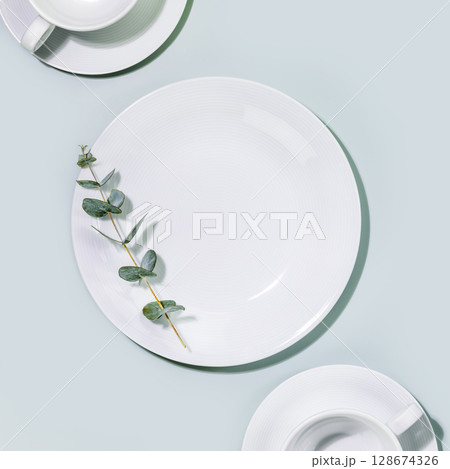 Clean, empty white dishes and cups with eucalyptus twig on blue background. Eco friendly washing dishes and cleaning. Dishes, porcelain background. Empty plate for your product placement. Flat lay 128674326