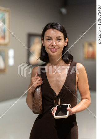 Woman with phone in hands in museum hall, view painting exhibition Woman with phone in hands in museum hall, view painting exhibition 128675467