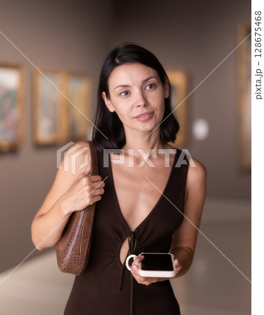 Woman with phone in hands in museum hall, view painting exhibition Woman with phone in hands in museum hall, view painting exhibition 128675468