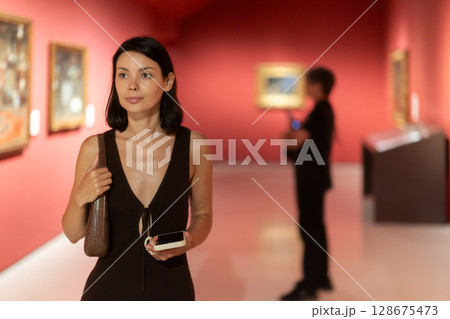 Woman with phone in hands in museum hall, view painting exhibition 128675473
