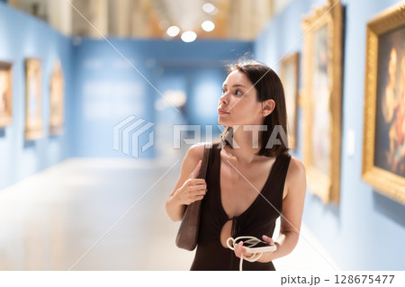 Woman with phone in hands in museum hall, view painting exhibition 128675477