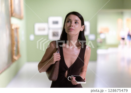 Woman with phone in hands in museum hall, view painting exhibition 128675479