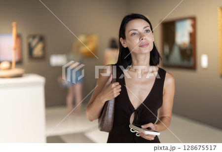 Woman with phone in hands in museum hall, view painting exhibition Woman with phone in hands in museum hall, view painting exhibition 128675517