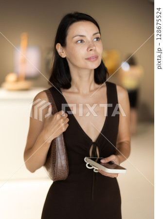 Woman with phone in hands in museum hall, view painting exhibition Woman with phone in hands in museum hall, view painting exhibition 128675524