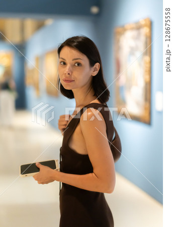 Woman with phone in hands in museum hall, view painting exhibition Woman with phone in hands in museum hall, view painting exhibition 128675558