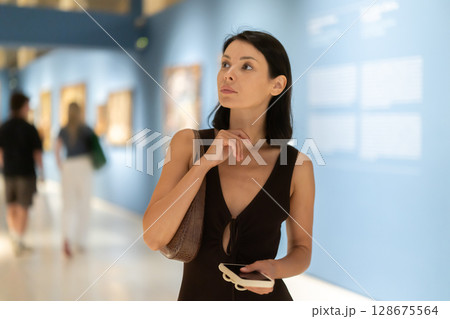 Woman with phone in hands in museum hall, view painting exhibition 128675564