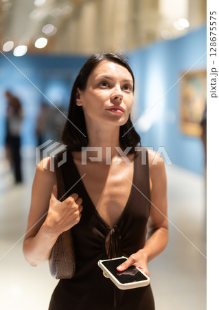 Woman with phone in hands in museum hall, view painting exhibition Woman with phone in hands in museum hall, view painting exhibition 128675575