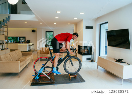 A triathlete riding a triathlon bike on a machine simulation in a modern living room A triathlete riding a triathlon bike on a machine simulation in a modern living room 128680571