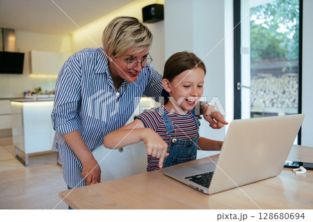 Mother with her daughter talking on laptop with family and friends while sitting in modern living room of big house. Mother with her daughter talking on laptop with family and friends while sitting in modern living room of big house. 128680694