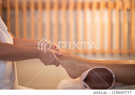 Foot Massage Therapy in a Relaxing Spa Setting. A professional massage therapist in a white uniform provides a soothing foot massage to a client in a calm and serene spa environment Foot Massage Therapy in a Relaxing Spa Setting. A professional massage therapist in a white uniform provides a soothing foot massage to a client in a calm and serene spa environment 128680737